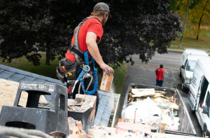 Operaio con imbracatura che rimuove materiali da un tetto durante lavori di ristrutturazione, con camion per detriti sottostante.