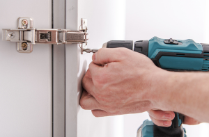 Mano che usa un avvitatore per montare una cerniera su uno sportello di mobile bianco.