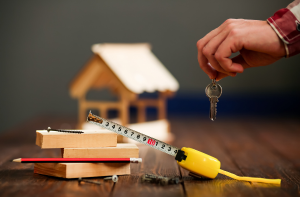 Mano che consegna una chiave con strumenti da lavoro e modellino di casa sul tavolo, simbolo di costruzione o vendita immobiliare.